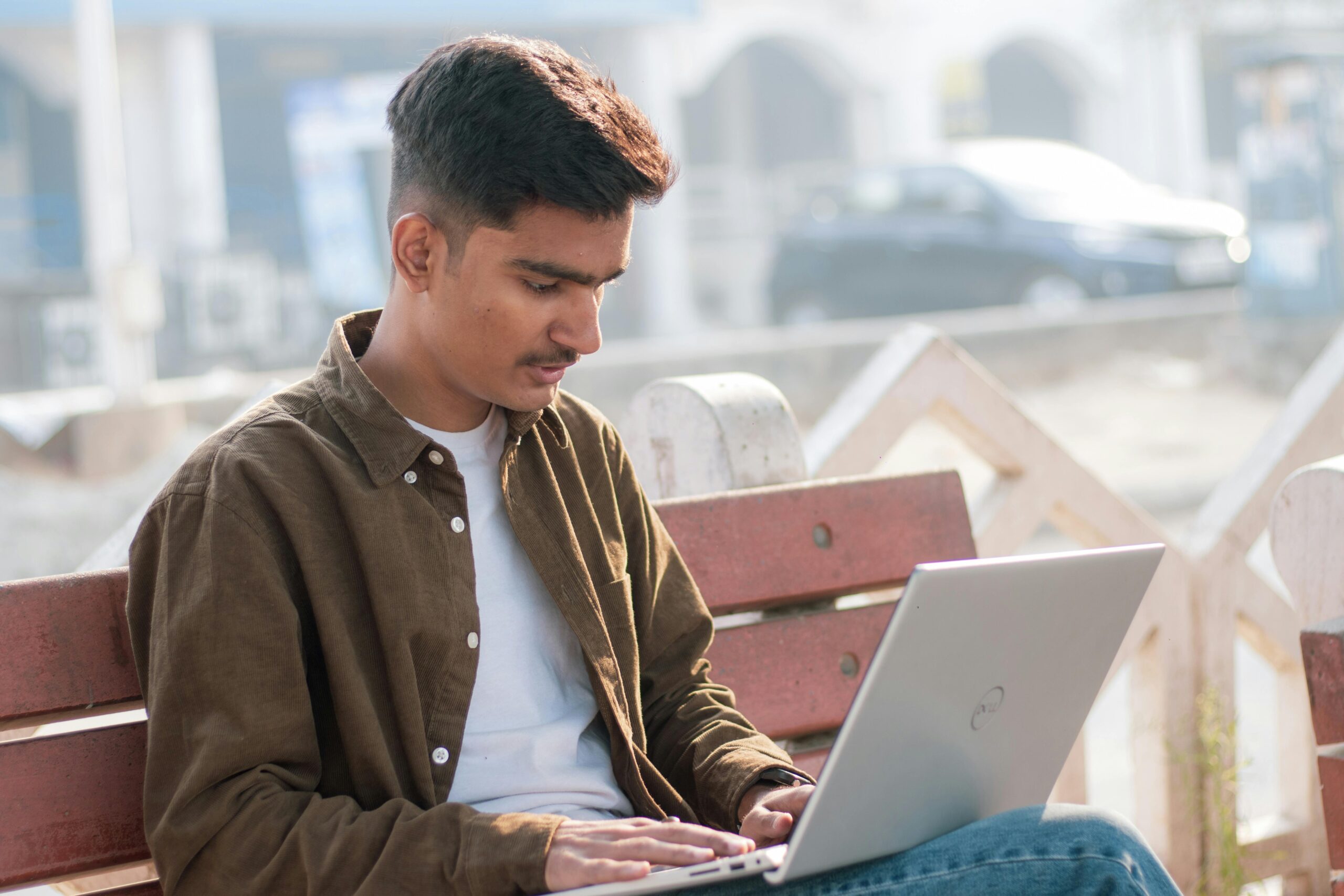 A young man focused on his laptop, working outdoors on a sunny day.